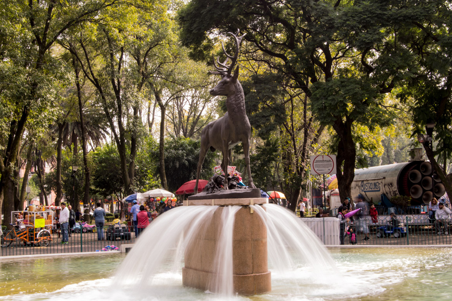 Fuente del parque de los venados, en el cual encima hay una estatua de un venado, en el fondo se aprecia a la gente disfruntando su día